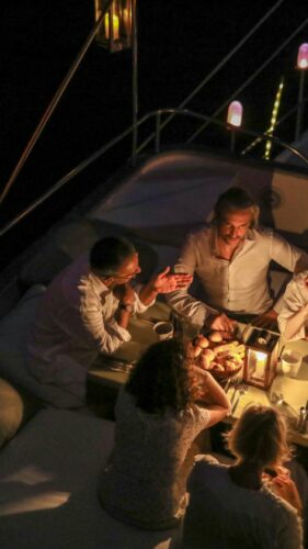 A group of six people, casually dressed, sit around a candlelit dinner table on a luxury yacht in the Maldives at night. Warm lighting from bulbs and candles creates a cozy ambiance as the group enjoys a meal and lively conversation.