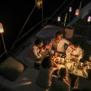A group of six people, casually dressed, sit around a candlelit dinner table on a luxury yacht in the Maldives at night. Warm lighting from bulbs and candles creates a cozy ambiance as the group enjoys a meal and lively conversation.