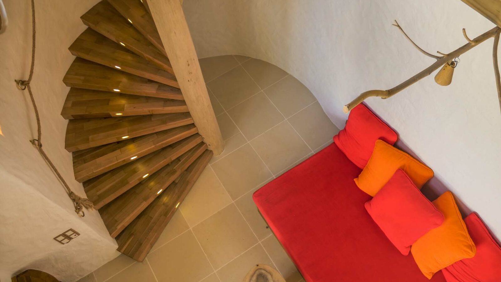 Top-down view of a cozy indoor space reminiscent of a Maldives luxury resort, featuring a curved wooden staircase with built-in lights ascending along a white wall. At the bottom, there's a red sofa accented with orange and red pillows, and a branch with a light fixture attached. Beige tiled floor underneath.