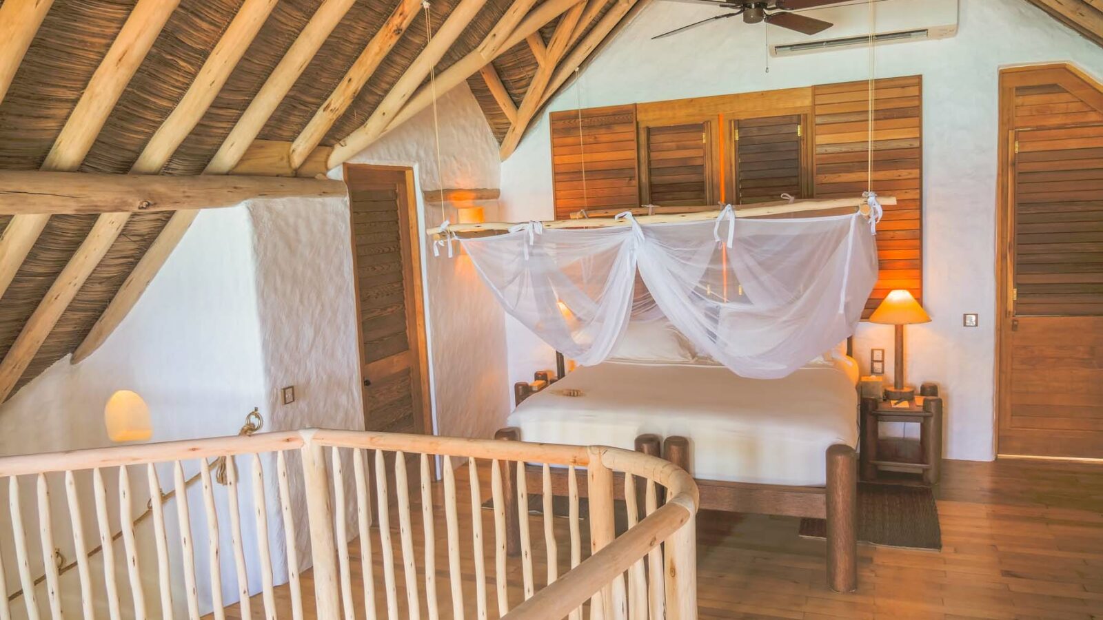 A cozy bedroom at Soneva Fushi Villas with a sloped wooden ceiling and exposed beams. The bed is covered with a white canopy mosquito net. There is a wooden railing in the foreground, and warm lighting from a lamp creates a serene ambiance. Experience Maldives luxury with wooden doors and window shutters completing the room.