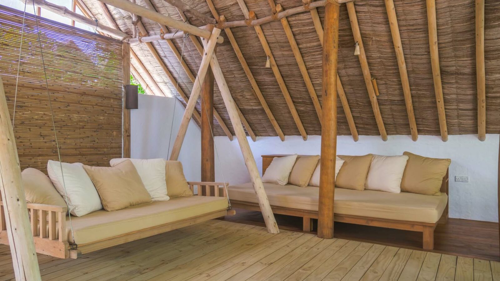 A cozy, rustic room reminiscent of a Maldives luxury resort, with a wooden floor and thatched roof, features two seating areas: a hanging daybed with white and beige cushions and a wooden sofa matching the same tone. Bamboo shades partially cover one wall, allowing diffused natural light to filter in.