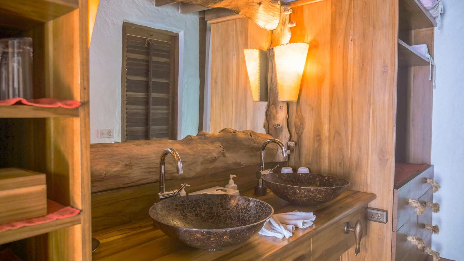 A rustic bathroom with wooden decor featuring a double sink setup with two metallic bowl sinks atop a wooden counter. Above the sinks are two artistic light fixtures that resemble tree trunks, reminiscent of the elegant design found in Maldives Luxury Resort Soneva Fushi Villas. The room includes a large mirror and wooden shelves for storage.