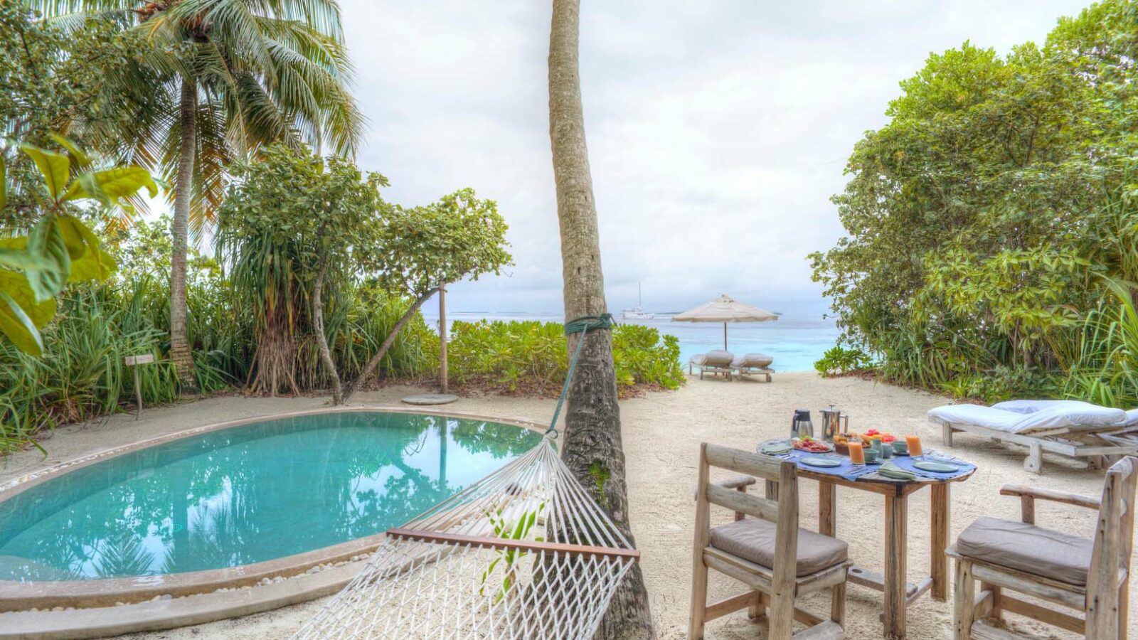 A tropical beach scene at the Maldives Luxury Resort features a hammock tied to a tree, a small round pool surrounded by foliage, and a table set with breakfast items. Two wooden chairs and a lounge chair with an umbrella are near the shoreline at Soneva Fushi Villas, with the ocean visible in the background.