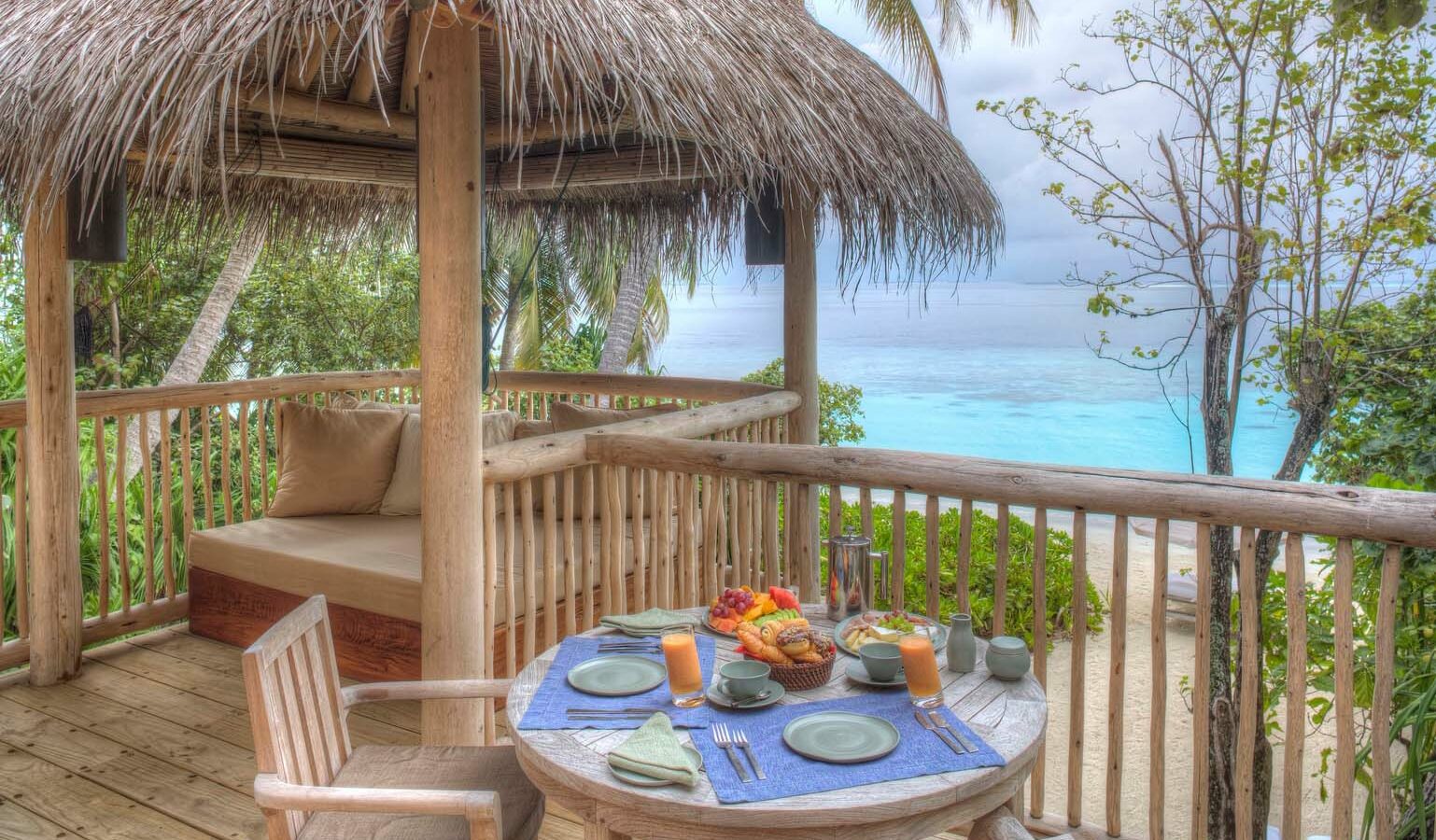 A tropical outdoor dining area at the Maldives' luxury resort, Soneva Fushi Villas, features a small round table set for two with plates, cups, fresh fruit, and glasses of juice. The seating area is under a thatched roof gazebo overlooking a serene beach and turquoise ocean, surrounded by lush greenery.
