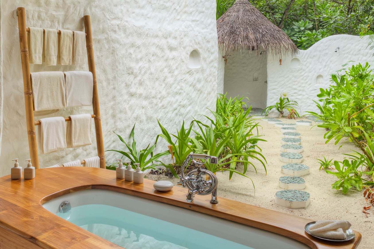 An outdoor bathroom at the Maldives Luxury Resort, Soneva Fushi Villas, features a wooden bathtub surrounded by lush greenery. Silver fixtures adorn the tub next to a white, textured wall with a towel rack. A pathway of rounded stones leads to the thatched-roof huts amidst tropical foliage.