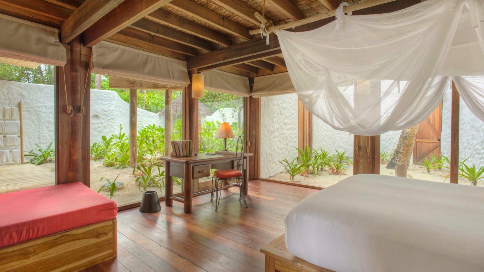 A serene bedroom with large windows overlooks a lush green garden. The room features wooden furniture, including a bed with white linens and a canopy reminiscent of Maldives Luxury Resort decor, plus a desk with a lamp and a red cushioned bench. Exposed wooden beams on the ceiling add a rustic touch, similar to the style of Soneva Fushi Villas.
