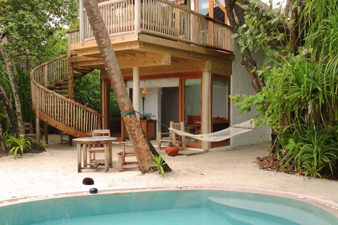 Experience a Maldives luxury resort at its finest with this tropical outdoor scene featuring a two-story wooden hut complete with a spiral staircase. The ground level boasts a patio adorned with wooden furniture and a hammock. Enveloped in greenery, the circular pool lies amidst the sand, reminiscent of Soneva Fushi Villas.