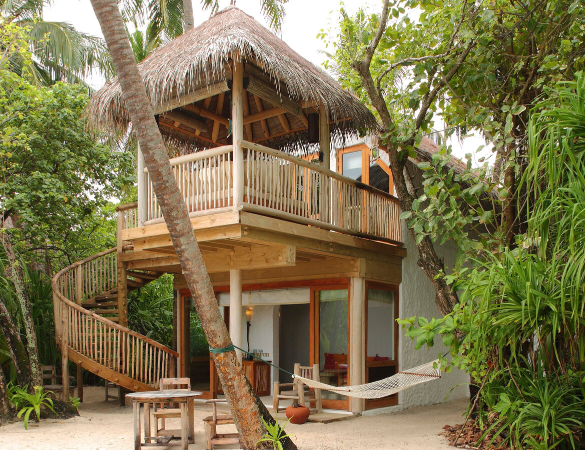 A two-story beach cottage made of wood and thatched roofing, nestled among tropical trees in a style reminiscent of the Maldives. The upper level boasts a balcony with railings and a palm frond roof. A hammock is strung between two trees, and there is a wooden table with benches on the sandy ground.