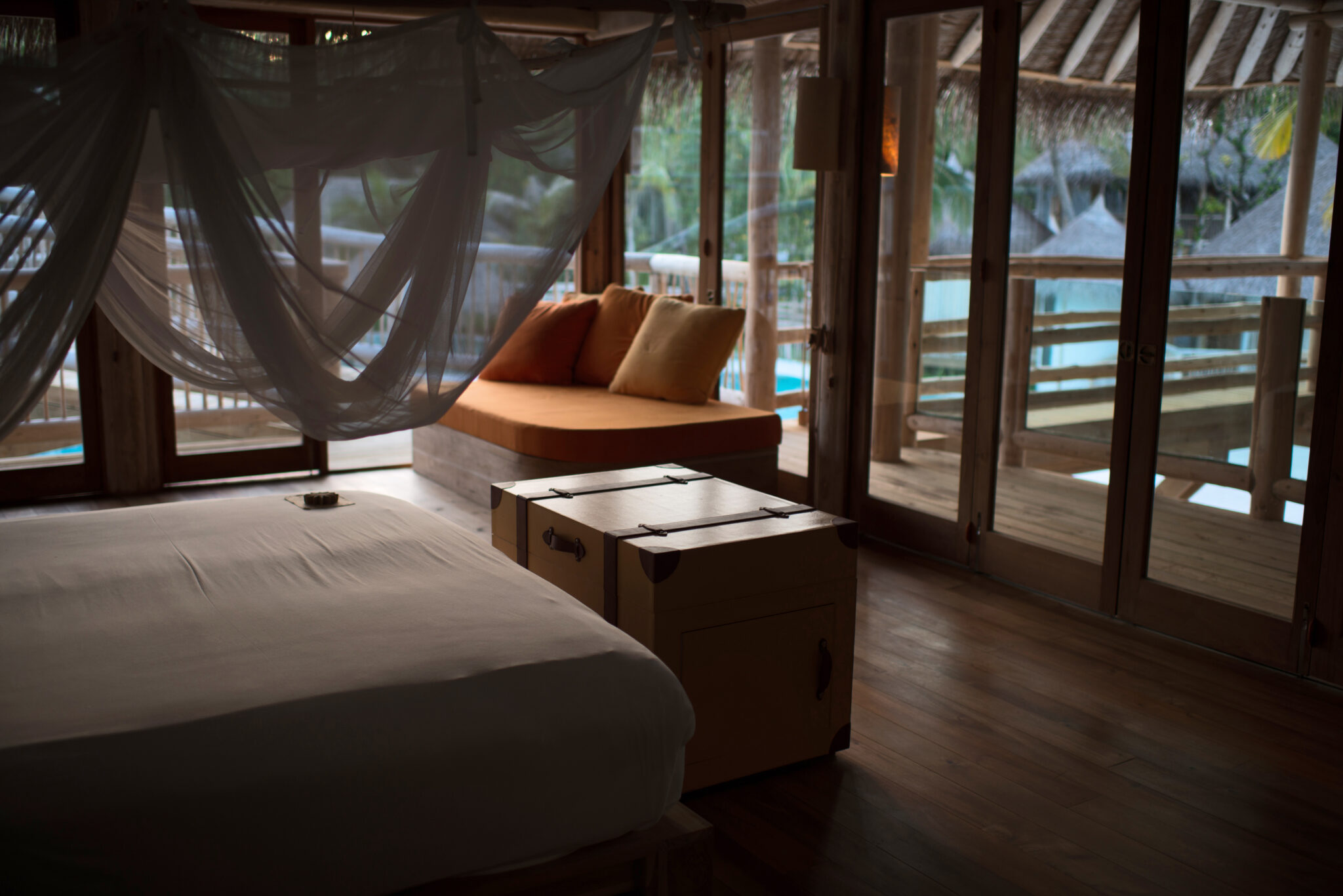 A serene Maldives villa interior featuring a bed with mosquito netting, a wooden chest at its foot, and an inviting cushioned daybed near large glass doors. The doors open to a deck with a view of a lush, tropical landscape. Warm, natural light fills the room, perfect for exclusive villa ownership.