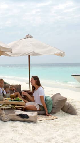 Four people sit on cushions under umbrellas, savoring a meal on a pristine beach. A table is set with food and drinks. In the background, the ocean is calm and a small boat is anchored near the shore. The sky is partly cloudy, enhancing this Maldives exclusive experience at Soneva with serene beauty.
