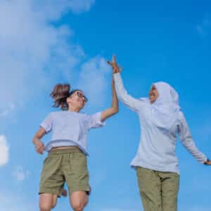 Two women in khaki shorts and white shirts, one wearing a hijab, are mid-jump high-fiving against a bright blue sky with a few clouds. Joyful with wide smiles, they seem to be celebrating the vibrant spirit of Maldives Soneva Namoona together.