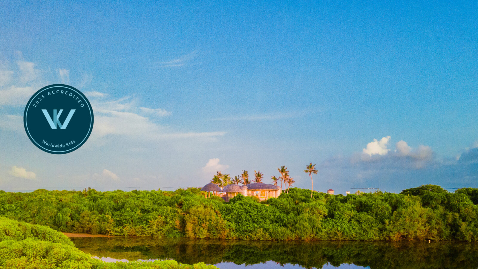 A scenic view of lush greenery with a building surrounded by trees in the distance evokes the serene charm of Soneva Jani. The sky is bright blue with scattered clouds, and a circular logo with the letter &quot;W&quot; graces the left side of this inviting family-friendly image.