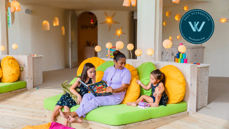 A woman sits on a lime green couch with two young girls, one with a book and the other a toy. In this vibrant Soneva Jani family room, colorful lights and toys surround them. A &quot;W Accredited Worldwide Kids&quot; badge is proudly displayed.