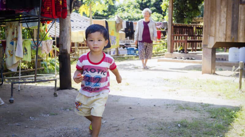 A young child in a red-striped shirt and yellow shorts walks on a dirt path toward the camera. Behind the child, an elderly woman stands near a wooden house with laundry hanging on a line. This scene, reminiscent of the serene landscapes supported by Maldives charity initiatives, features lush greenery and trees.