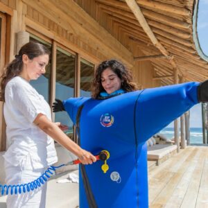 A woman is helping another person fully dressed in a blue inflatable suit with extended arms on a wooden deck near the beach. The woman operates an air tube connected to the inflatable suit, evoking a quirky scene at a Maldives spa. In the background, there's a wooden house and spiral staircase.