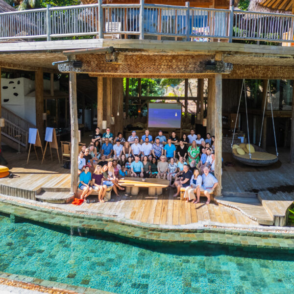 A large group of people from the Salt Group gathered on a wooden deck near a pool, in front of a rustic, multi-level building. Some sit on benches and chairs, while others stand. The setting is tropical, with lush greenery surrounding the Soneva-inspired area.