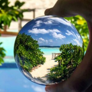 A clear spherical lens is held up against a beach backdrop, showing a magnified, inverted view of a sandy pathway leading to a turquoise sea with lush greenery on either side and a chair at the end. The scene evokes memories of a serene family holiday in Maldives&#039; Soneva resort, surrounded by more greenery and a bright sky.