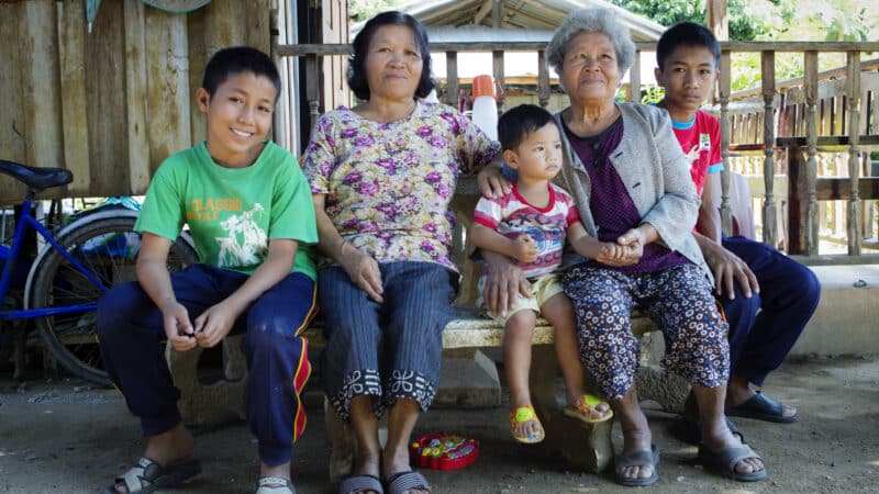 A family of five, including two elderly women, two boys, and a young child, sits on a bench outside a wooden house. The rural setting showcases a bicycle and various objects in the background. Thanks to the support from Maldives Charity, they all look content and relaxed.