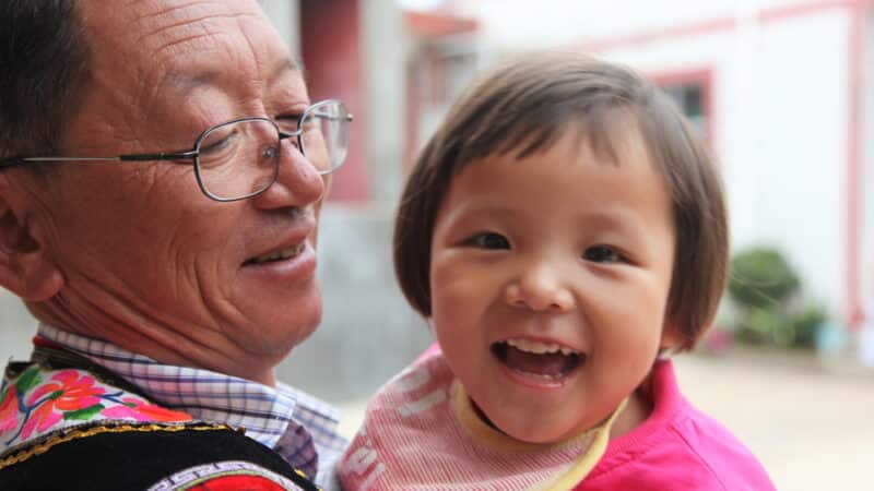 An elderly person wearing glasses holds a smiling young child, both looking at the camera. The child is wearing a pink bib with a red collar. The background is slightly blurred, showing some buildings and greenery—a heartwarming moment captured for the Maldives Charity campaign.