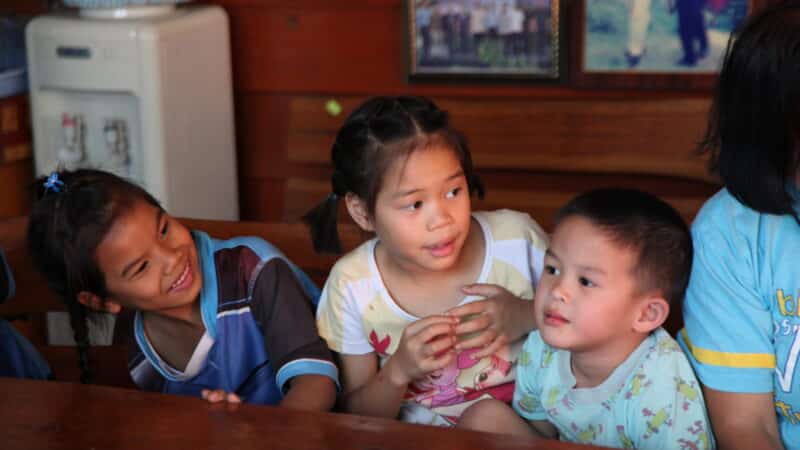 Three children are sitting at a wooden table indoors, gently smiling and looking in different directions. They appear engaged in conversation or a shared activity. The background includes framed pictures from a Maldives charity hanging on a wooden wall and a water dispenser on the left.