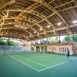 An indoor tennis court with a wooden arched ceiling. One player is on the court, ready to hit a ball, while several people sit on colorful bean bags to the left. The background features tropical plants and ample natural light filtering through the structure, reminiscent of Soneva Exclusive Offers' tranquil settings.