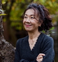 A woman with dark hair and a navy sweater stands outdoors with arms crossed. She is smiling and looking upwards. The background features a tree and blurred greenery.