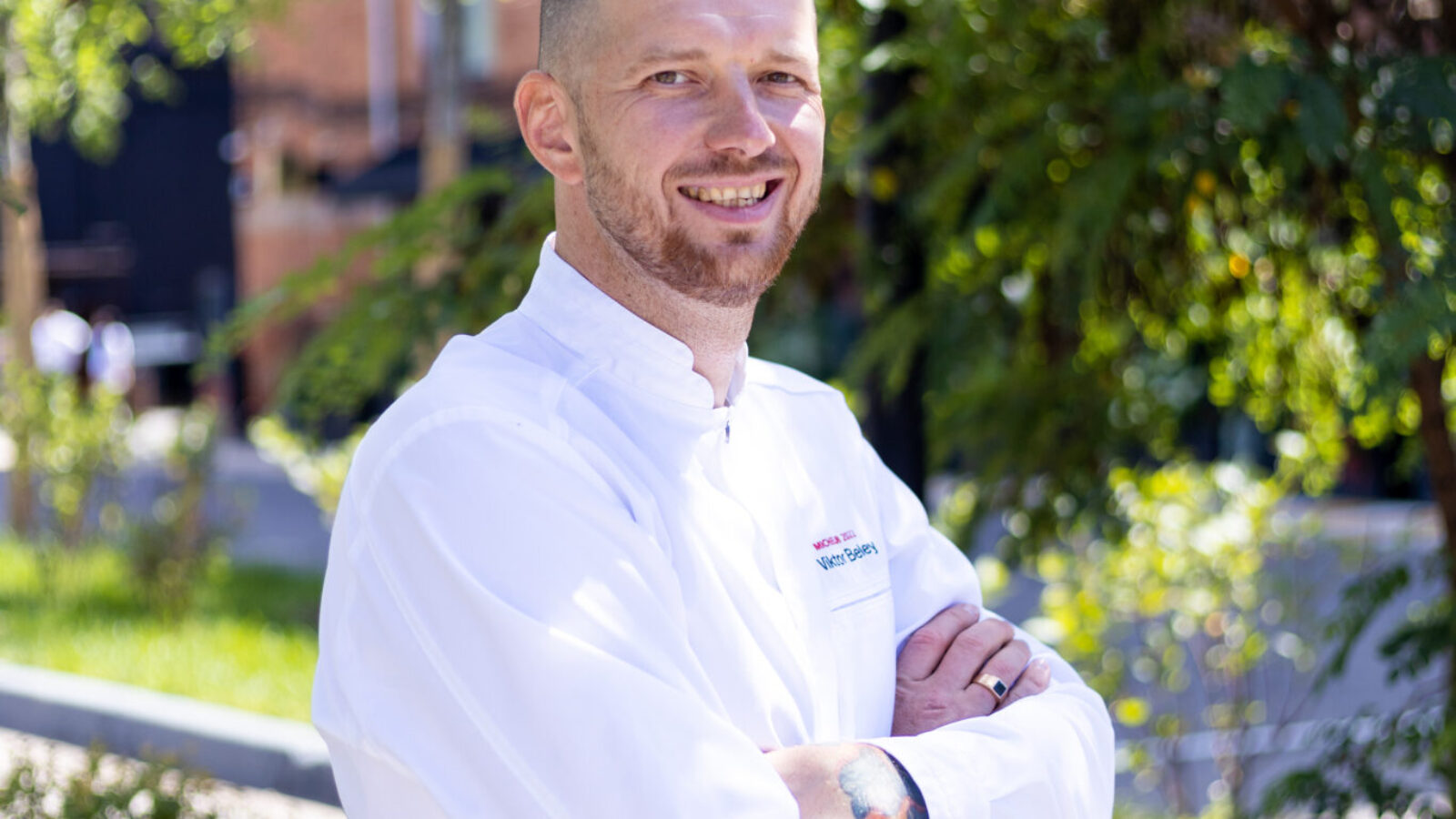 A person wearing a white chef&#039;s coat stands outdoors with arms crossed, smiling. They are in a garden setting with lush greenery in the background and sunlight filtering through the leaves.