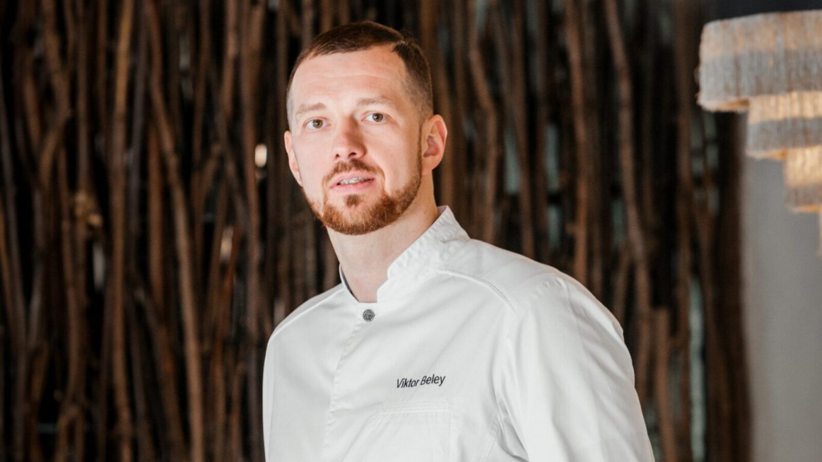 A man with a beard in a white chef&#039;s jacket stands confidently with hands in pockets. He is in a restaurant with modern decor, featuring a wooden backdrop and dining tables. The lighting is warm and ambient.