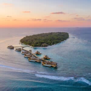 Aerial view of a tropical island at sunset, featuring dense greenery and luxurious over-water villas. The villas, available for Maldives villa ownership, are connected by a wooden walkway extending into the turquoise ocean. The sky displays hues of orange, pink, and purple, reflecting on the calm waters.