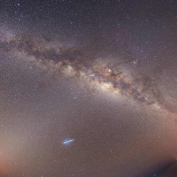 银河系横跨夜空，无数繁星点缀其间，画面令人震撼。近处可见一颗流星，远处隐约可见地平线和一些建筑物的灯光，营造出一种类似于马尔代夫 Soneva 酒店夜晚的独特体验。
