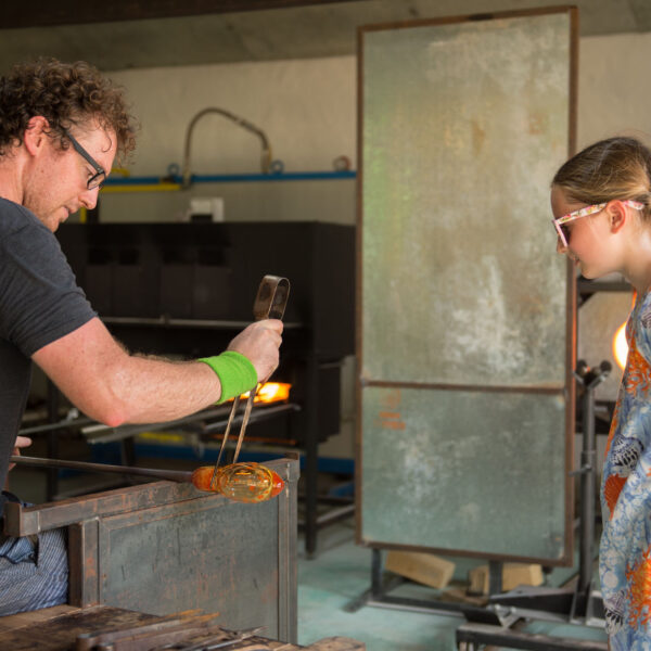 A male glassblower with curly hair demonstrates his craft to a young girl with braided hair and wearing a colorful dress. Using glassblowing tools, he creates intricate designs while she watches attentively, experiencing this Maldives exclusive experience at Soneva's renowned workshop.