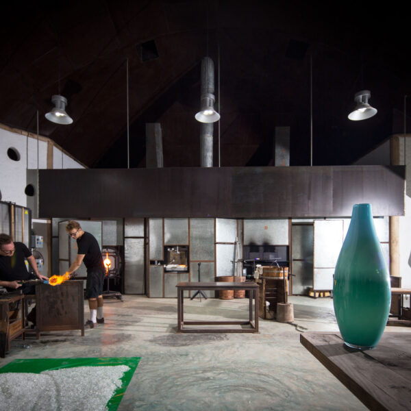 A spacious, dimly-lit glassblowing studio with high ceilings. Two people are working with molten glass near a furnace. A large green vase sits prominently on a wooden table in the foreground, reminiscent of an exclusive Soneva design. Various glassblowing tools and equipment are scattered throughout the space.