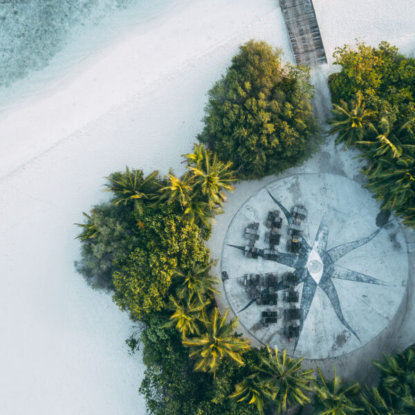 Aerial view of a circular beachside seating area surrounded by lush greenery and palm trees. The white sandy beach and turquoise waters are visible to the left, with a wooden pathway leading to the seating area from the top right corner—an exclusive Maldives experience reminiscent of Soneva&#039;s luxurious retreats.