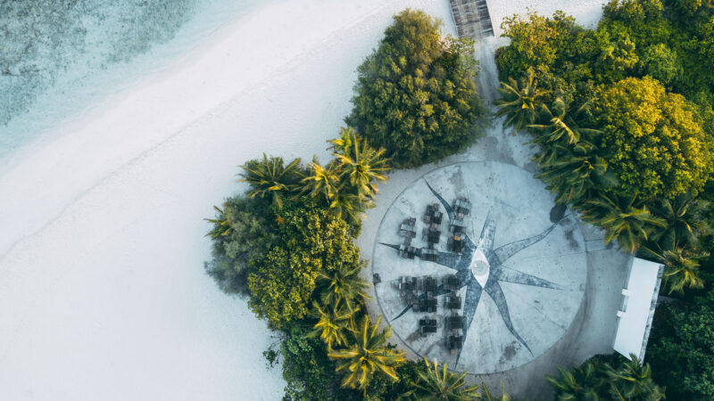 Aerial view of a circular beachside seating area surrounded by lush greenery and palm trees. The white sandy beach and turquoise waters are visible to the left, with a wooden pathway leading to the seating area from the top right corner—an exclusive Maldives experience reminiscent of Soneva&#039;s luxurious retreats.