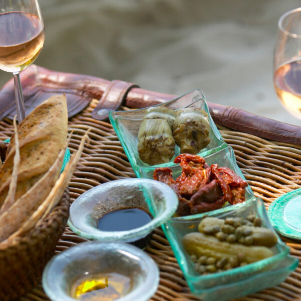 A wicker tray on the beach holds two glasses of rosé wine, a basket of sliced bread, dishes of artichokes, sun-dried tomatoes, and pickled vegetables. Several small bowls contain oil and balsamic vinegar for dipping – the perfect setup for a Maldives Exclusive Experience courtesy of Soneva.