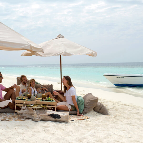 A group of four, two men and two women, enjoy a meal on a sandy beach under large umbrellas as part of their Maldives Exclusive Experience. They sit on cushions around a low table with food and drink. A blue and white boat is anchored nearby in the calm turquoise water. The sky is partly cloudy.