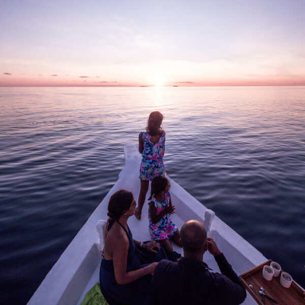 A family of five sits at the front of a boat, watching a sunset over a calm ocean. Two adults hold their children, while another person is seated nearby. A dolphin is seen leaping out of the water to the right. The sky is painted with shades of pink and orange, epitomizing a Maldives exclusive experience by Soneva.