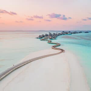 Aerial view of a winding wooden pathway stretching across a sandy beach towards overwater bungalows with the turquoise sea on both sides. The sky, painted with soft hues of pink and orange during sunset, creates a serene and picturesque scene perfect for a Soneva Maldives holiday.