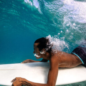 A surfer with short hair and wearing striped shorts is underwater, holding onto his surfboard as a wave passes over him. The turquoise water, part of the Maldives exclusive experience at Soneva, is clear, showing bubbles around the surfer&#039;s face and body.