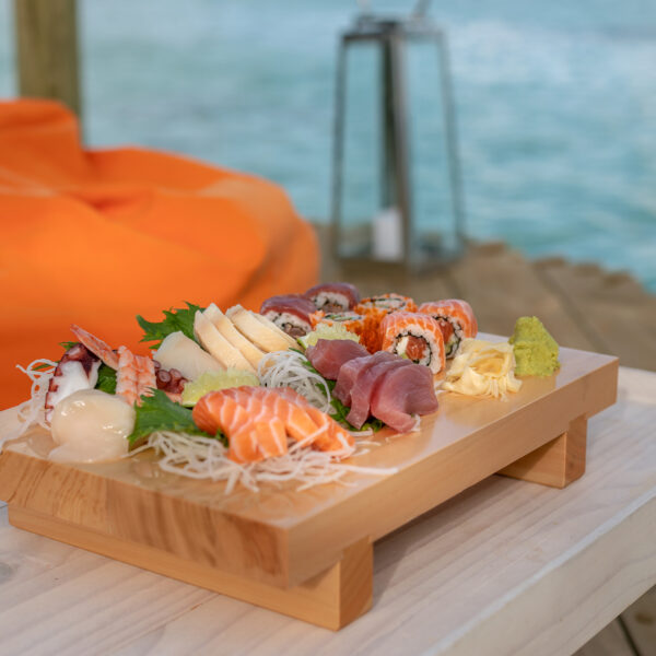 A wooden tray filled with assorted sushi, sashimi, and garnishes sits on a light wooden table. An orange bean bag and a blurred view of a body of water are visible in the background, suggesting a relaxed, outdoor dining setting that&#039;s part of the exclusive Soneva experience in the Maldives.