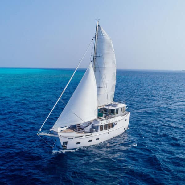 A white sailboat glides on clear blue ocean waters under a bright sky, epitomizing a Maldives luxury yacht holiday. White sails are fully unfurled, catching the wind, as the horizon stretches into the distance where the vibrant blue sea meets the lighter blue sky.