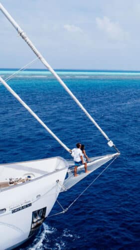 A person stands at the bow of a sailboat cruising through the deep blue waters under a clear Maldivian sky. They are holding onto the rigging. A yellow lifeboat is visible on the deck. The horizon shows a strip of turquoise water, indicating a nearby reef, embodying the essence of a Soneva exclusive experience.