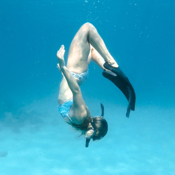 A person wearing a snorkel, mask, and swim fins is underwater, gracefully curved in a backward dive position. The blue ocean surrounds them as they enjoy an exclusive diving experience, reminiscent of Thailand&#039;s clear waters and luxurious retreats like Soneva.