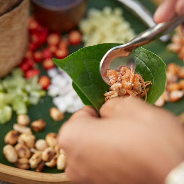 一个人用勺子将包括坚果在内的切碎配料混合物放在一片叶子上的特写。叶子被放在一个装有更多坚果、红辣椒、切碎的蔬菜和编织容器的托盘上。这项泰国独有的体验捕捉到了 Soneva 烹饪艺术的精髓。