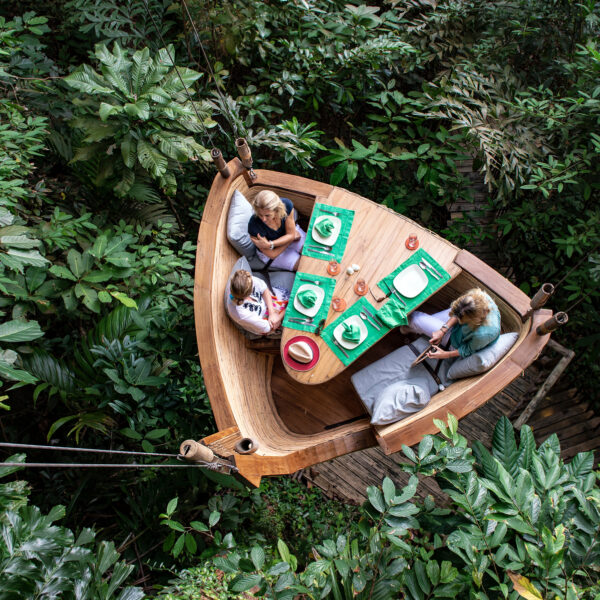 Three people dining at a wooden table shaped like a boat, suspended high among dense green foliage. The table is set with plates, glasses, and napkins. The diners sit on cushioned bench seating, surrounded by lush tropical vegetation—an exclusive Soneva experience in Thailand.