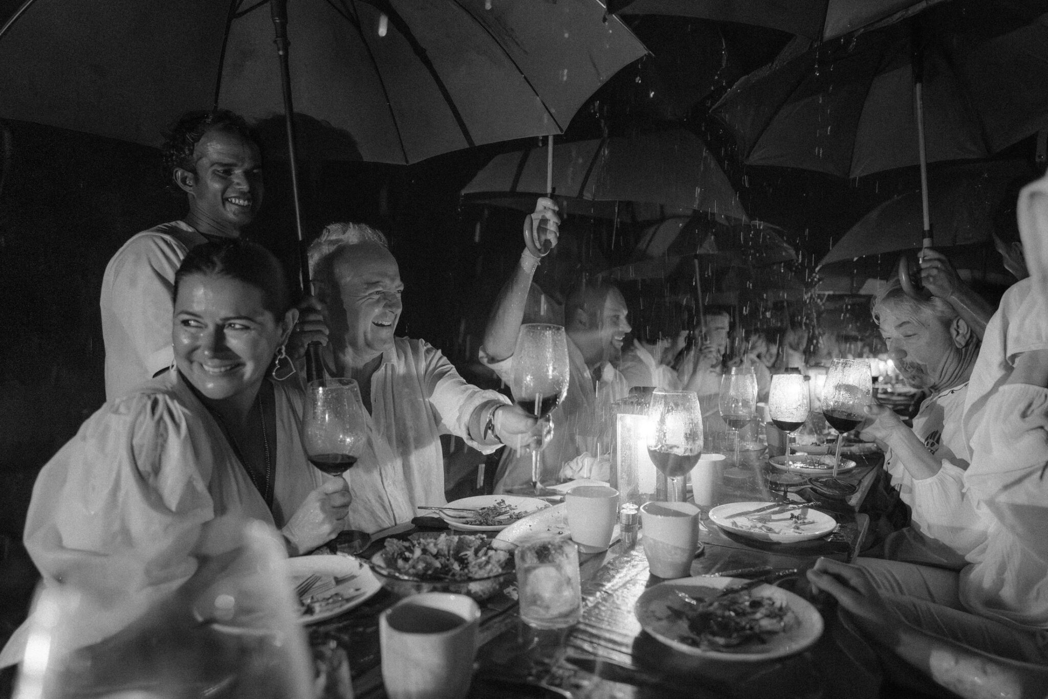 A black-and-white photo features a group of smiling diners seated at an outdoor table, holding umbrellas above their heads to shield from the rain. Candlelight illuminates the scene, reminiscent of the serene ambiance one might find soaking in wellness at Soneva Soul.