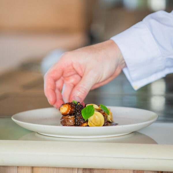 A hand carefully places a garnish on a gourmet dish in a kitchen. The plate contains a small, artfully arranged selection of food items. The background shows a blurred kitchen setting.
