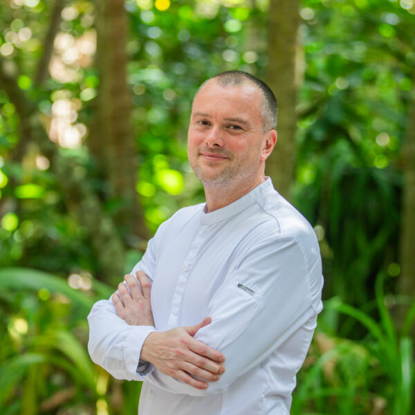 A person in a white chef's jacket stands with arms crossed, smiling in an outdoor setting. The background features lush green foliage and trees.