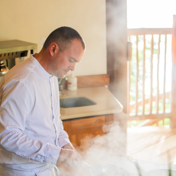 A chef in a white uniform stands by a stove, cooking amidst rising steam. Sunlight filters through a wooden railing in the background, illuminating the kitchen area.