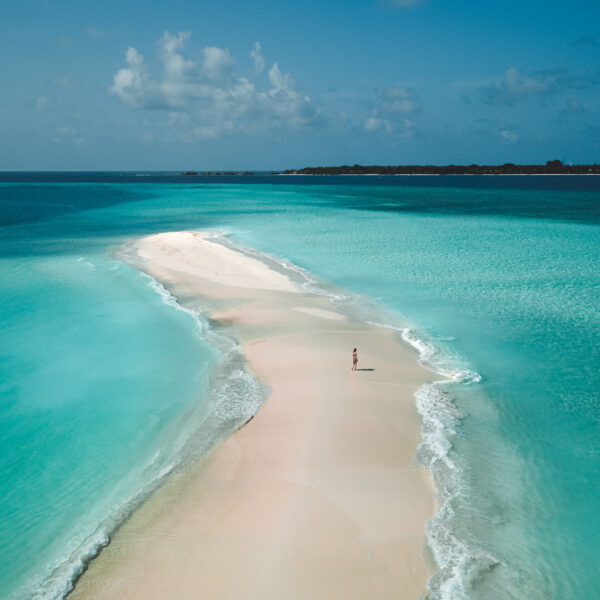 A lone person walks along a narrow sandbar surrounded by crystal-clear turquoise waters on both sides under a clear blue sky with a few scattered clouds. The sandbar stretches far into the distance, creating a serene and picturesque tropical scene reminiscent of the Maldives' luxury resorts like Soneva Fushi Villas.
